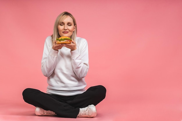 Ritratto di giovane bella donna affamata che mangia il panino del croissant. Ritratto isolato di donna con fast food su sfondo rosa. Concetto di dieta. Seduto sul pavimento nella posa del loto.