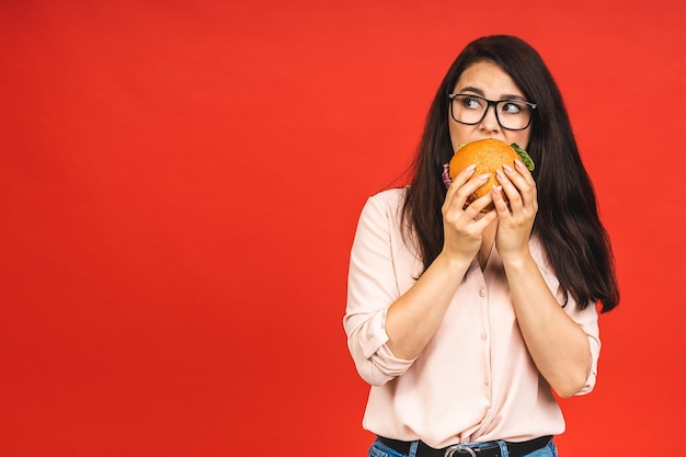 Ritratto di giovane bella donna affamata mangiare hamburger Ritratto isolato di studente con fast food su sfondo rosso Concetto di dieta