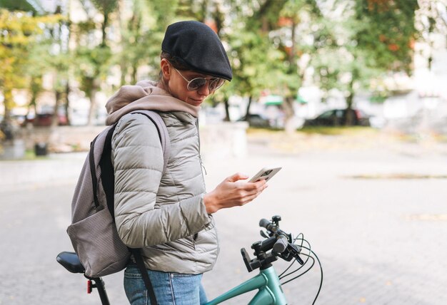 Ritratto di giovane bella donna alla moda in berretto e occhiali da sole utilizzando il cellulare in bicicletta in una soleggiata giornata autunnale nel parco cittadino