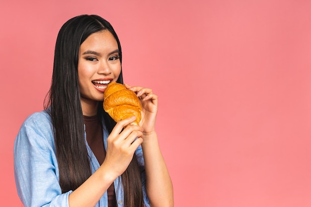 Ritratto di giovane bella donna asiatica affamata mangiare croissant Ritratto isolato di donna con fast food su sfondo rosa Concetto di colazione dietetica