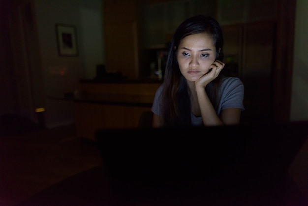 Ritratto di giovane bella donna asiatica utilizzando laptop nel buio soggiorno