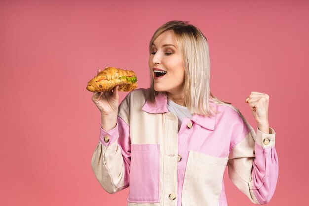 Ritratto di giovane bella donna bionda affamata che mangia il panino del croissant. Ritratto isolato di donna con fast food su sfondo rosa. Concetto di dieta.