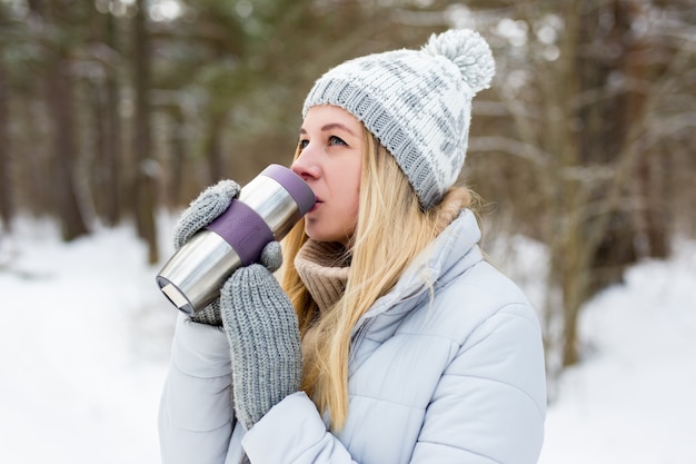 Ritratto di giovane bella donna bionda che beve caffè nel parco invernale o nella foresta