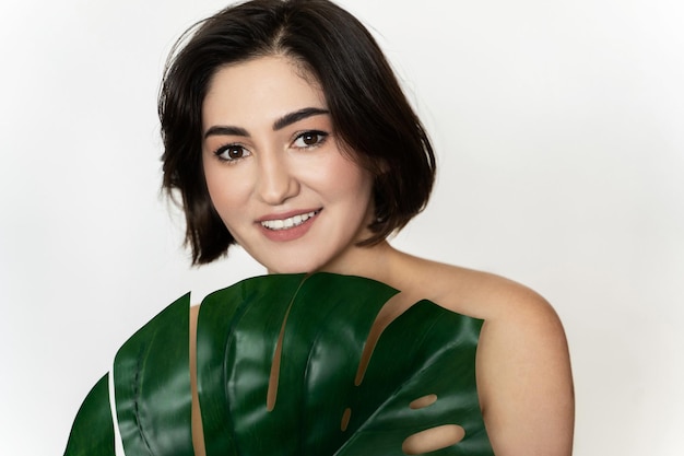 Ritratto di giovane bella donna bruna con una foglia tropicale di monstera deliziosa su sfondo bianco