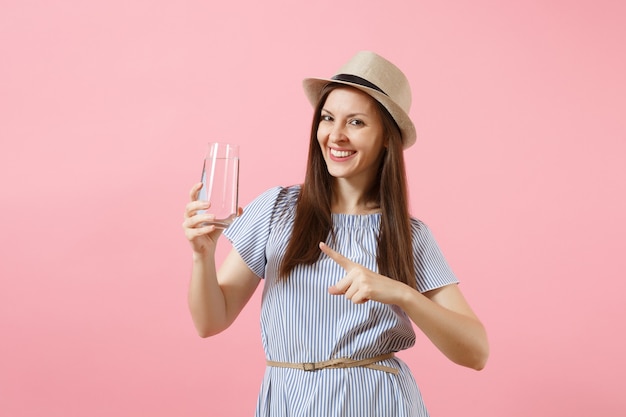 Ritratto di giovane bella donna in abito blu, cappello che tiene e beve acqua pura fresca e limpida dal vetro isolato su sfondo rosa. Stile di vita sano, persone, concetto di emozioni sincere. Copia spazio