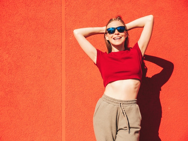 Ritratto di giovane bella donna sorridente hipster in abiti estivi alla moda Donna spensierata sexy in posa vicino al muro rosso in strada al tramonto Modello positivo all'apertoFelice e allegro