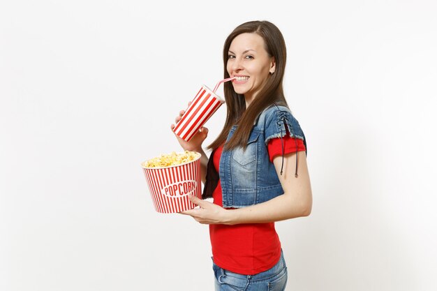 Ritratto di giovane bella donna sorridente in abiti casual guardando film, tenendo secchio di popcorn, bevendo da una tazza di plastica di soda o cola isolato su sfondo bianco. Emozioni al cinema.