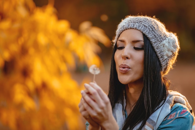 Ritratto di giovane bella donna sorridente positiva che soffia dente di leone nel parco autunnale.