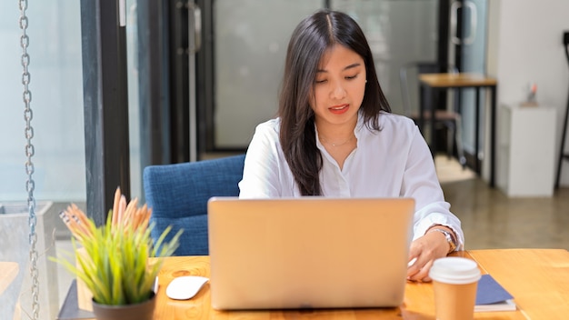 Ritratto di giovane bella femmina in camicia bianca che lavora con il computer portatile nella comoda stanza dell'ufficio