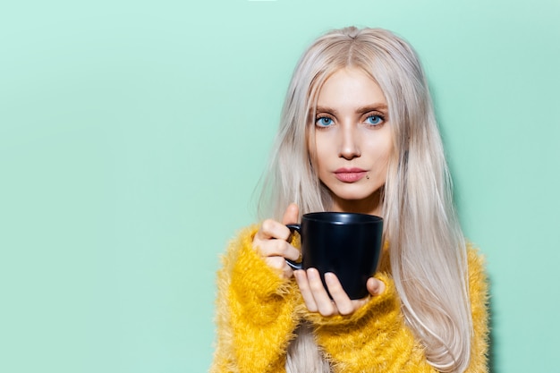 Ritratto di giovane bella ragazza carina con gli occhi azzurri, che tiene in mano una tazza di ceramica nera su uno sfondo di colore verde pastello con spazio per le copie. Indossa un maglione giallo.