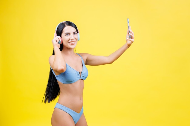 Ritratto di giovane bella ragazza con cuffie wireless sulla testa prendendo selfie per i social network sorridendo e ridendo della fotocamera del telefono isolata su sfondo giallo