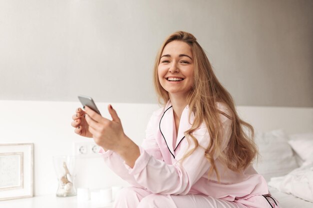 Ritratto di giovane bella signora seduta sul letto con il cellulare in mano e felicemente guardando a porte chiuse a casa