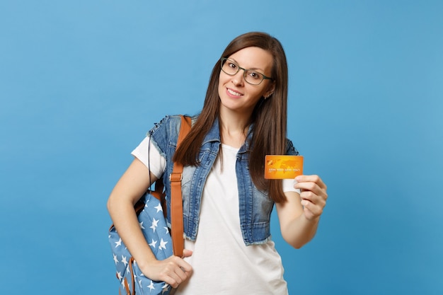 Ritratto di giovane bella studentessa interessata piacevole in vestiti di denim, occhiali con zaino tenere la carta di credito isolata su sfondo blu. Istruzione nel concetto di college universitario di liceo.