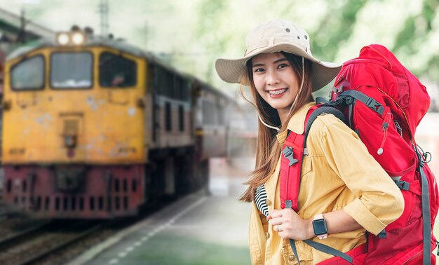 Ritratto di giovane bella viaggiatrice con zaino in attesa della partenza alla stazione ferroviaria, sorridente e guardando davanti