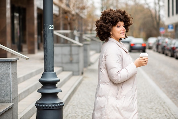 Ritratto di giovane, carina donna caucasica di 40 anni, abbigliamento casual sportivo, camminando agitando la mano, tazza di caffè usa e getta. Acconciatura afro. Gente allegra, vita di città