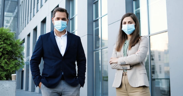 Ritratto di giovane caucasico uomo e donna in stile business e maschere mediche in piedi a distanza sociale e guardando la fotocamera. Uomo d'affari e donna d'affari all'aperto durante la pandemia.