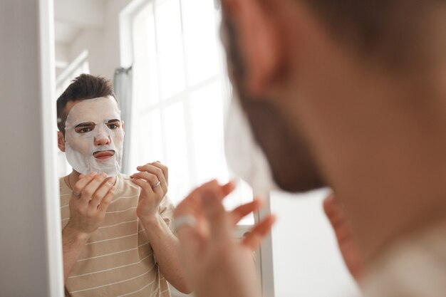 Ritratto di giovane che applica la maschera facciale mentre si guarda allo specchio durante la routine mattutina per la cura della pelle, copia spazio