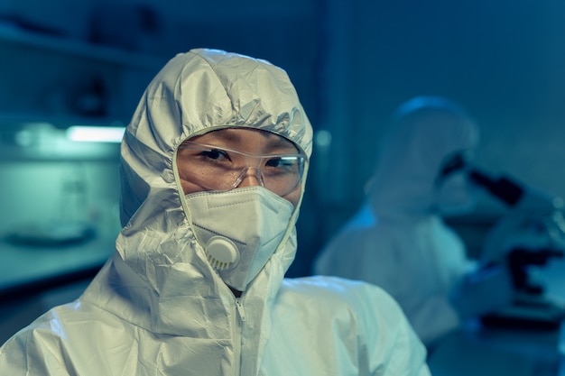 Ritratto di giovane chimico asiatico in abiti da lavoro protettivi e occhiali che guardano la telecamera mentre si lavora in laboratorio