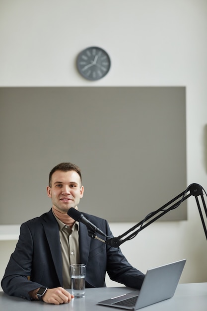 Ritratto di giovane conduttore radiofonico sorridente davanti mentre parla al microfono al tavolo con il computer portatile