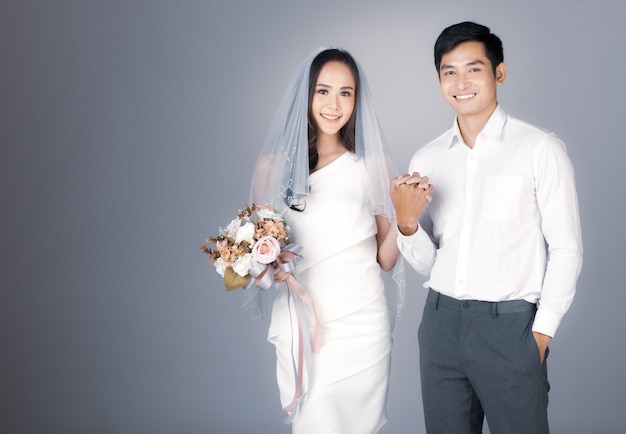 Ritratto di giovane coppia asiatica attraente che si tiene per mano, uomo che indossa una camicia bianca, donna che indossa abito da sposa con velo che tiene un mazzo di fiori. Concetto per la fotografia prematrimoniale.
