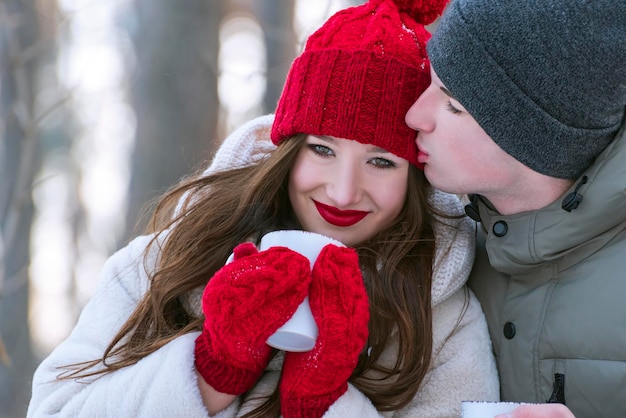 Ritratto di giovane coppia di innamorati in inverno all'aperto. La ragazza in cappello rosso e guanti tiene la tazza.