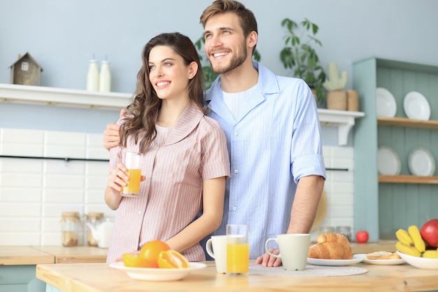 Ritratto di giovane coppia felice in pigiama che cucina insieme in cucina, bevendo succo d'arancia al mattino a casa.