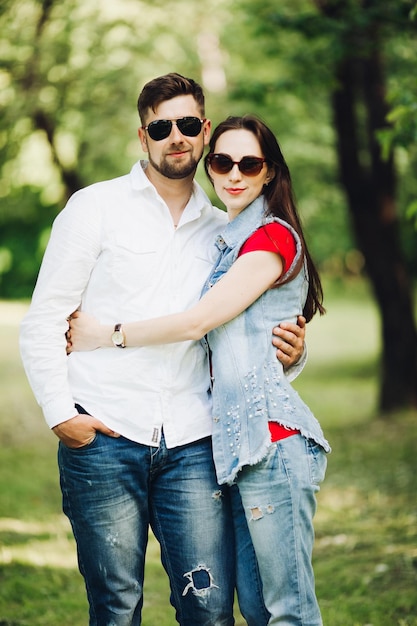 Ritratto di giovane coppia felice innamorata sorridente e abbracciata in giardino Dolci amanti che indossano eleganti occhiali da sole scuri e abiti casual in posa e guardando la fotocamera durante il giorno nel parco estivo