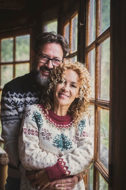 Ritratto di giovane coppia matura sorridente e in piedi per una foto Uomo e donna che indossano un maglione caldo con decorazioni natalizie e colori Persone innamorate in uno chalet di legno o a casa nel tempo libero