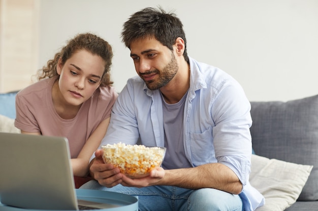 Ritratto di giovane coppia moderna guardando film online tramite laptop e tenendo una grande ciotola di pop corn