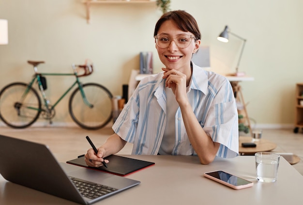 Ritratto di giovane designer femminile che sorride alla telecamera e utilizza la tavoletta con penna sul posto di lavoro dell'ufficio a casa