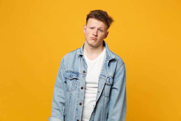 Ritratto di giovane dispiaciuto in abiti casual denim in piedi e guardando la fotocamera isolata su sfondo giallo arancione parete in studio. Persone sincere emozioni concetto di stile di vita. Mock up spazio di copia.