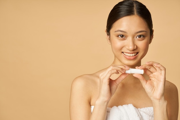 Ritratto di giovane donna adorabile avvolta in un asciugamano sorridente mentre si tiene il balsamo per le labbra in posa isolata