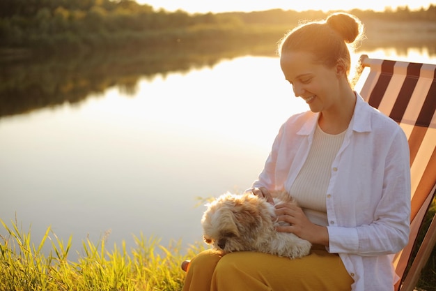 Ritratto di giovane donna adulta caucasica sorridente che indossa una camicia bianca e il suo cane pechinese seduto al tramonto vicino al fiume che accarezza il suo animale preferito