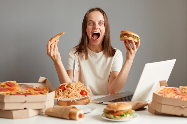 Ritratto di giovane donna adulta eccitata che mangia fast food mentre guarda un film sul computer portatile seduto al tavolo contro il muro grigio con in mano un panino e una fetta di pizza urlando