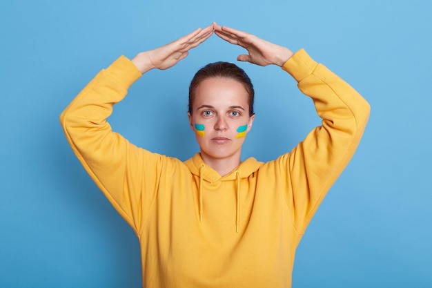Ritratto di giovane donna adulta in felpa con cappuccio gialla con bandiera ucraina sulle guance tenendo le mani sopra la testa fa cadere il gesto del tetto in cassaforte nella sua patria in posa isolata su sfondo blu