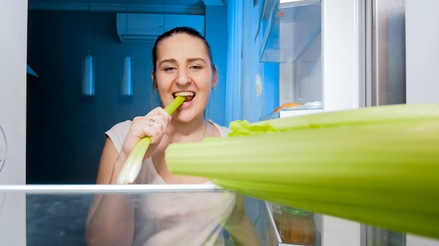 Ritratto di giovane donna affamata in piedi al frigorifero aperto e mangia sedano sano di notte. Concetto di dieta e alimentazione sana.