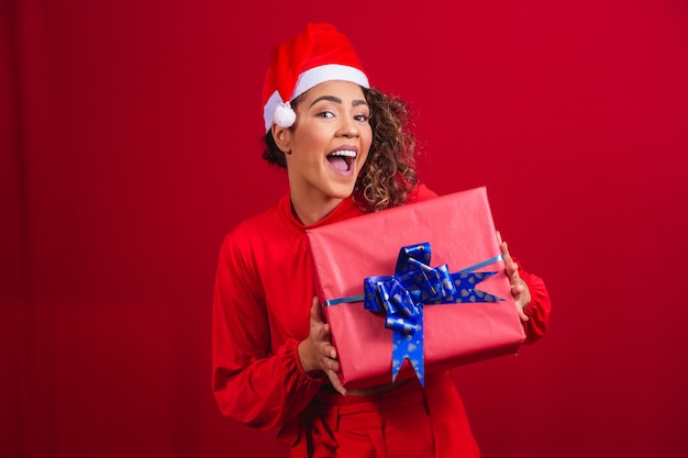 Ritratto di giovane donna afro in cappello di babbo natale che tiene un regalo su sfondo rosso. concetto di notte di natale