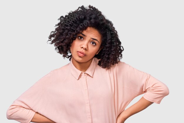 Ritratto di giovane donna afro interrogata che guarda seria alla telecamera durante la discussione vestita di camicia rosa con capelli ricci in posa in studio La ragazza afroamericana studentesca ha un'espressione confusa