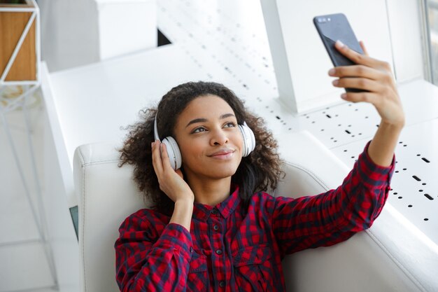 Ritratto di giovane donna afroamericana che indossa le cuffie prendendo selfie foto sul cellulare in cafe