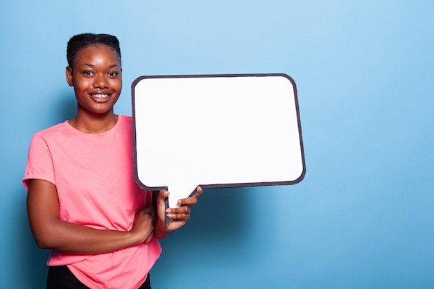 Ritratto di giovane donna afroamericana che sorride alla macchina fotografica mentre tiene il banner bianco del quiz in piedi in studio con sfondo blu. Concetto di pubblicità. Concetto di lavagna