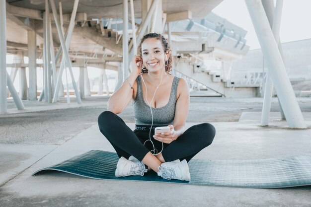 Ritratto di giovane donna afroamericana sorridente che si riposa mentre ascolta musica dopo l'allenamento. Ascolto di musica su smartphone. Sport urbano .Leggings abbigliamento e top. Copia spazio tappetino yoga