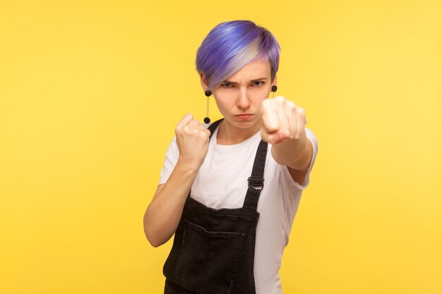 Ritratto di giovane donna aggressiva hipster con capelli corti tinti viola in tuta di jeans che punzona davanti alla telecamera che mostra gesto di boxe autodifesa isolata su sfondo giallo studio shot