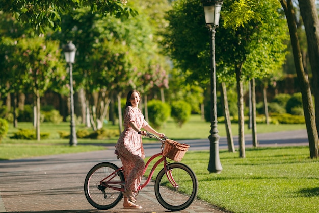 Ritratto di giovane donna alla moda in abito floreale lungo rosa ferma a cavalcare bici d'epoca con cesto per acquisti, cibo o fiori all'aperto, splendido tempo di ricreazione femminile nel parco primaverile o estivo.