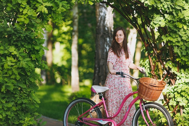 Ritratto di giovane donna alla moda in abito floreale lungo rosa ferma a cavalcare sotto l'arco di quercia in bicicletta con cesto per acquisti, cibo o fiori all'aperto, tempo di ricreazione femminile carino nel parco primaverile o estivo