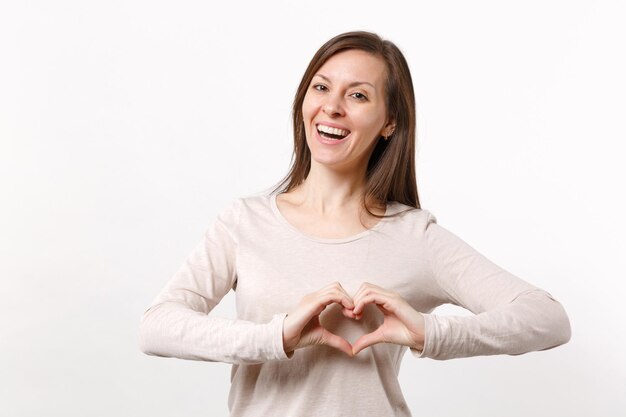 Ritratto di giovane donna allegra che ride sorridente in abiti leggeri che mostrano il cuore di forma con le mani isolate sul fondo bianco della parete. Persone sincere emozioni, concetto di stile di vita. Mock up spazio di copia.