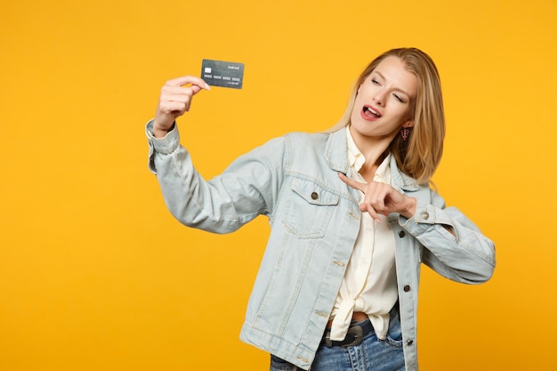 Ritratto di giovane donna allegra in abiti casual denim che punta il dito indice sulla carta di credito isolata su sfondo giallo arancione parete in studio. Concetto di stile di vita delle persone. Mock up spazio di copia.