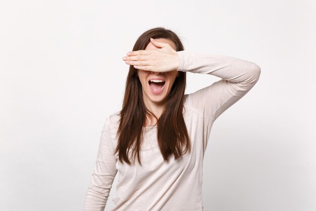 Ritratto di giovane donna allegra in abiti leggeri che coprono gli occhi con la mano che tiene la bocca aperta isolata su sfondo bianco della parete in studio. Persone sincere emozioni concetto di stile di vita. Mock up spazio di copia.