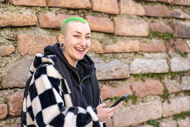 Ritratto di giovane donna androgina autentica con taglio di capelli corto e donna lesbica sorridente genuina di colore verde che utilizza smartphone ragazza felice con piercing e codice di abbigliamento street fashion