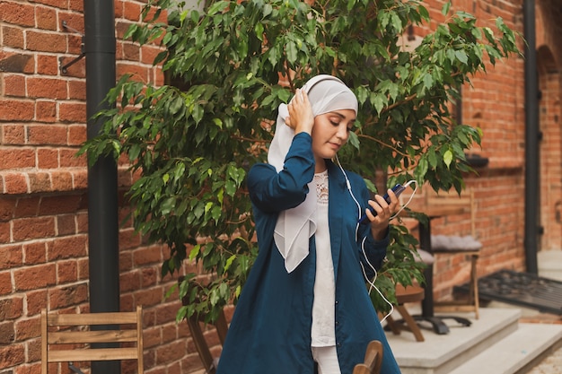 Ritratto di giovane donna araba moderna che tiene il telefono cellulare e ascolta la musica in cuffia.