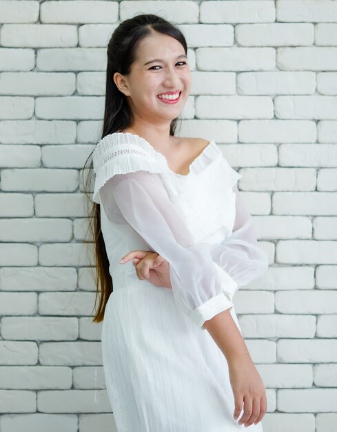 Ritratto di giovane donna asiatica adolescente in abito bianco in piedi sullo sfondo di un muro di mattoni bianchi con posa sorridente sicura di sé e positiva.